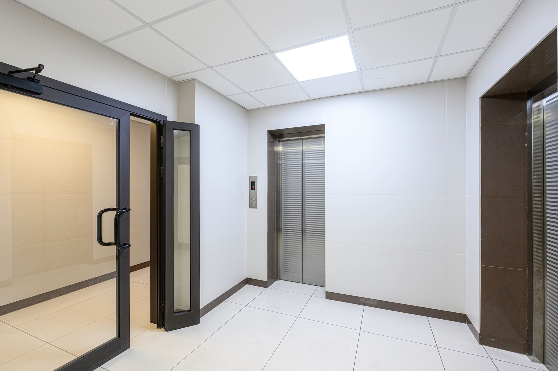 clean, modern interior space featuring white tiled walls and floor. An elevator and an open glass door are visible. The room is well-lit, giving it a bright and spacious feel